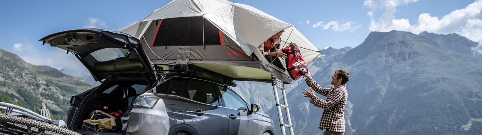 A couple set-up their roof tent on top of their vehicle in front of a mountain background